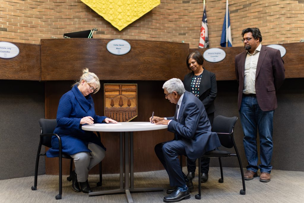 Radhe sitting at table signing a donation cheque while Krishna and Rohit are standing behind and watching with smiles.